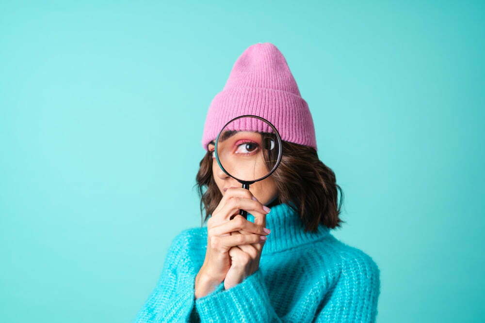 cozy portrait young woman knitted blue sweater pink hat with bright makeup holding magnifying glass fooling around having fun Taking Power Into Your Own Hands: How to Spot Greenwashing in 2023